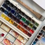 A vibrant watercolor paint set with a wooden paintbrush on a desk.